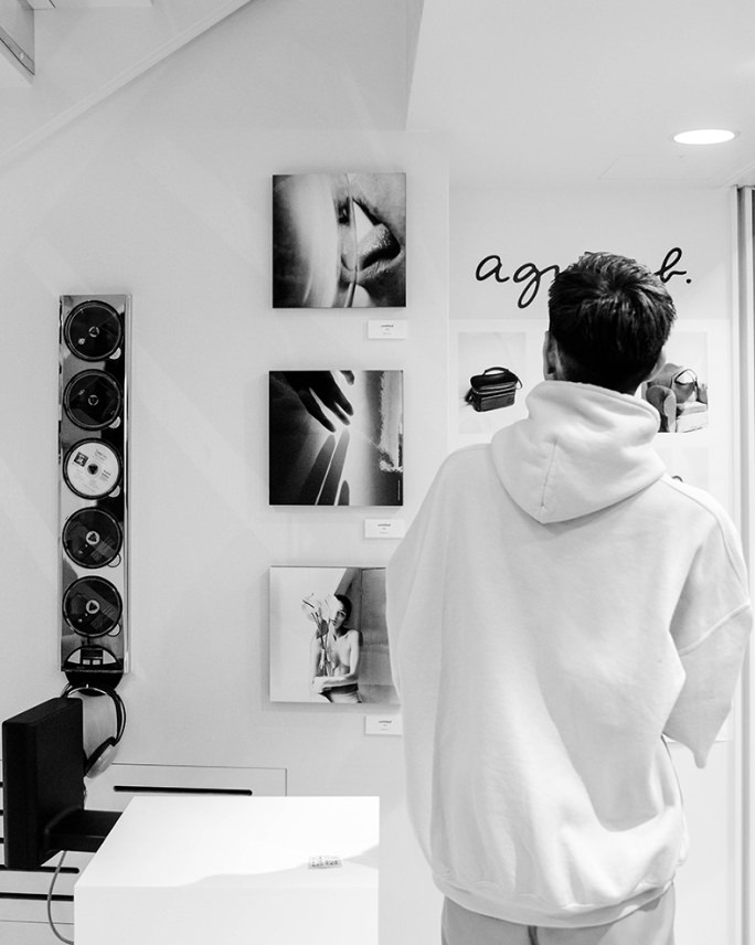 「agnès b.」のロゴがある白い壁に飾られた3枚のモノクロ写真作品を、白いフーディーを着た男性が背後から眺めている。