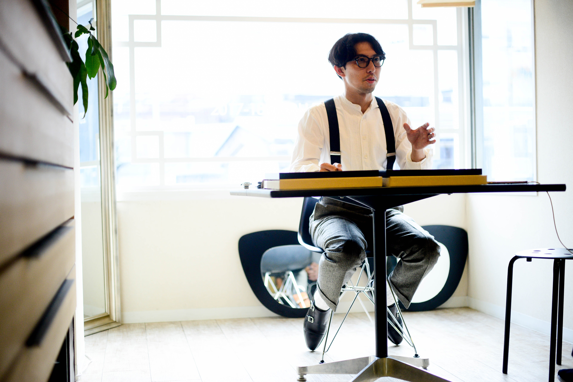 明るい窓際のテーブルで話す今泉悠。テーブルの上には黄色い縁取りのファイルが置かれている。